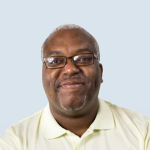 Gary is a dark-skinned man. He's wearing a yellow shirt with glasses and has a big smile on his face. He has short gray hair and brown eyes. He is pictured against a blue-grey background.