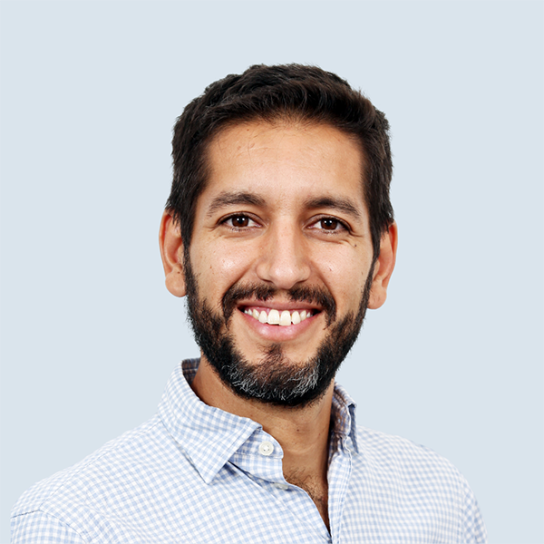 Marcelo is a medium-skinned man with short dark brown hair, a dark brown beard and brown eyes. He is wearing a blue and white checkered shirt. He is pictured smiling broadly against a blue-grey background.