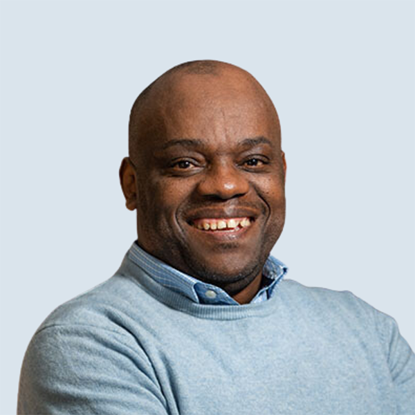 A portrait of Samkelo Blom, a dark-skinned man, with a bright and welcoming smile. He wears a blue cardigan over a shirt, radiating confidence and enthusiasm. He is pictured in front of a blue-grey background.