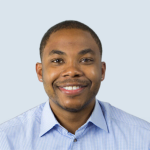 Mitch is a brown-skinned male with brown eyes and short black hair. He has a short brown beard and is wearing a blue and white checkered shirt. He is pictured smiling energetically against a blue-grey background.