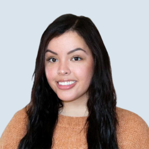 Natalie is a light-medium/medium-skinned female smiling softly. She has long, dark brown/black wavy hair and is wearing a soft orange wool knit sweater. She is pictured against a blue-grey background.