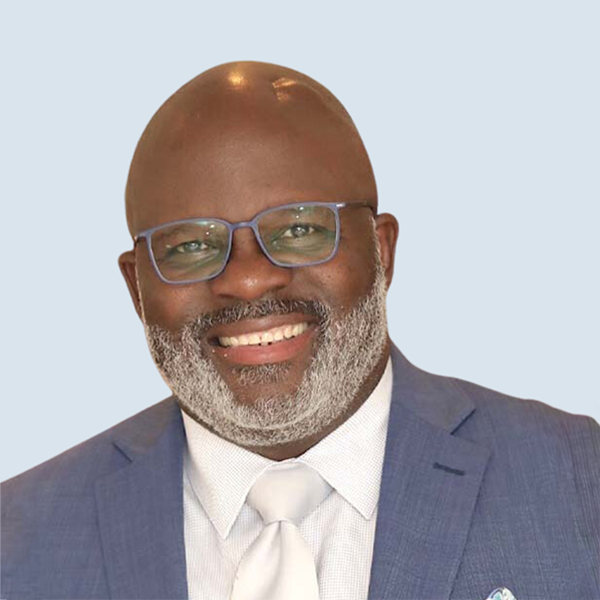 Doug Harris is an African American man with a bald head and grey beard. Doug is wearing a light blue suit paired with a crisp white dress shirt and a white silk tie, he completes his look with grey-framed glasses, adding a touch of sophistication. He is smiling broadly against a blue-grey background.