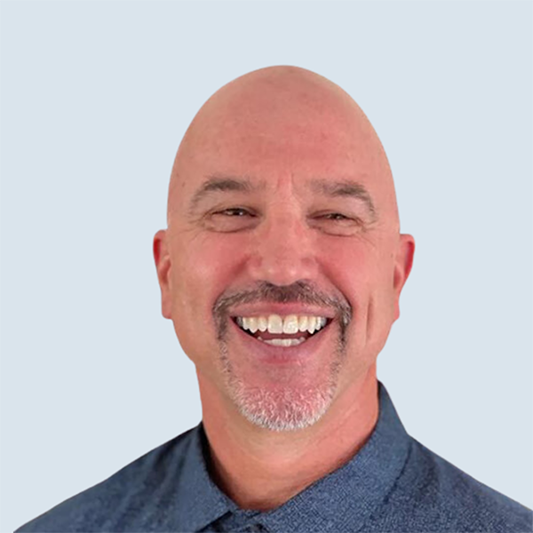 Brian is a bald, white, guy with a salt and pepper gray goatee that is crisply maintained. He is also wearing a navy blue, long sleeved dress shirt. He is pictured with a big smile on his face after someone told a joke, in front of a blue-grey background.