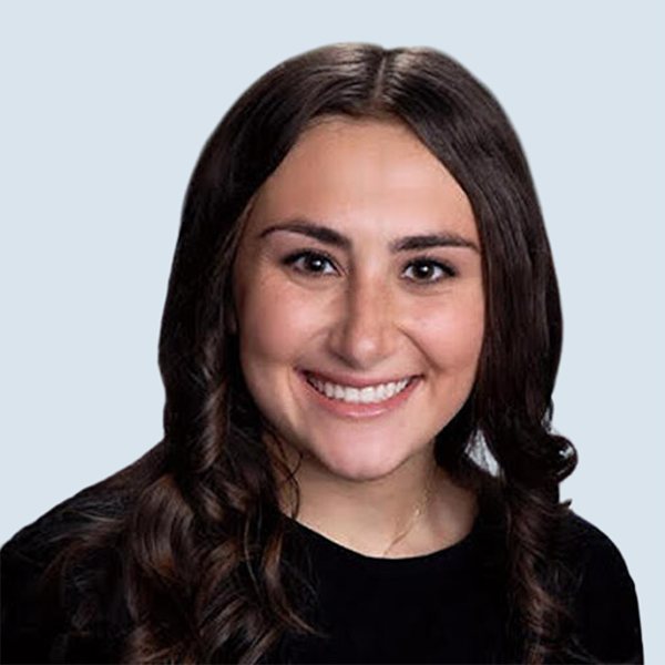 Arielle is a light-skinned woman with dark brown eyes and long brown hair. She is wearing a black shirt and is smiling into the camera against a blue-grey background.