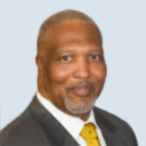 Jessie is a medium-dark skinned man with short gray hair and a gray goatee. He is wearing a black suit jacket, a white collared shirt and a yellow tie. He is pictured in front of a grey-blue background.