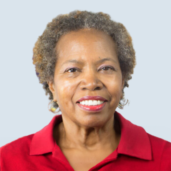 Cherè is a medium brown skinned woman with mingled grey short curly hair. She is wearing a red collared shirt. She has a bright smile and is pictured on a blue-grey background.