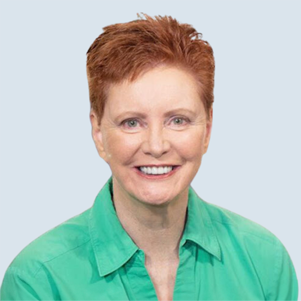 Sue is a light-skinned woman with short red hair and green eyes. She is wearing a green collared shirt. She is pictured smiling broadly aginst a blue-grey background.