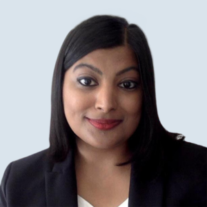 Fariha is a medium dark skinned female smiling. She has long black hair and is wearing a black blazer with a white shirt. She is pictured against a blue-grey background.