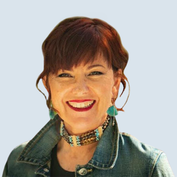 Hanlie is a freckled, light-skinned female human. She has short, wavy hair and is wearing a denim jacket, a choker and turquoise earrings. She is looking straight at the camera sporting a broad smile against a blue-grey background.
